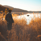 A person stands in a field of tall grass looking out over a calm lake at sunrise.