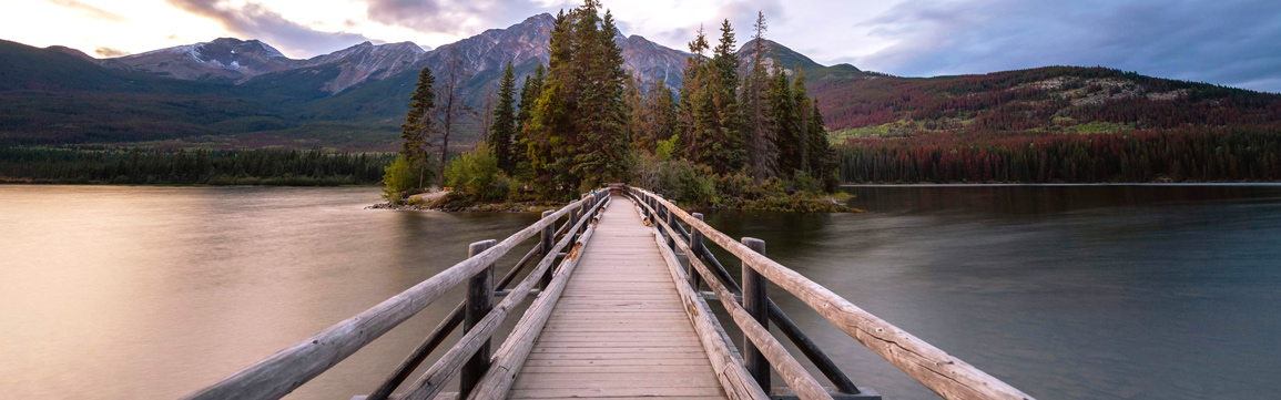 A balanced, symmetrical view of a wooden bridge leads across a calm lake to a small, pine-covered island, with large mountains rising in the background.