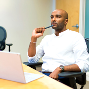 Man sitting in an office looking up thoughtfully from a laptop, with a pen in his hand.